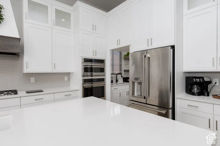 Kitchen featuring white shaker-style cabinetry with glass-front uppers, an expansive solid-surface island, stainless steel built-in double wall ovens, a matching french-door refrigerator, and a neutral subway tile backsplash