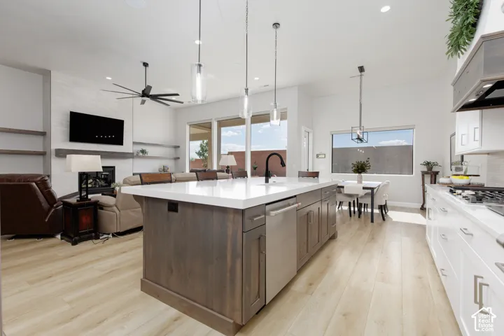 Kitchen featuring a large central island with light-toned countertop, custom wood-finish cabinetry, stainless steel dishwasher, modern pendant lighting, and light-toned wood-finish flooring