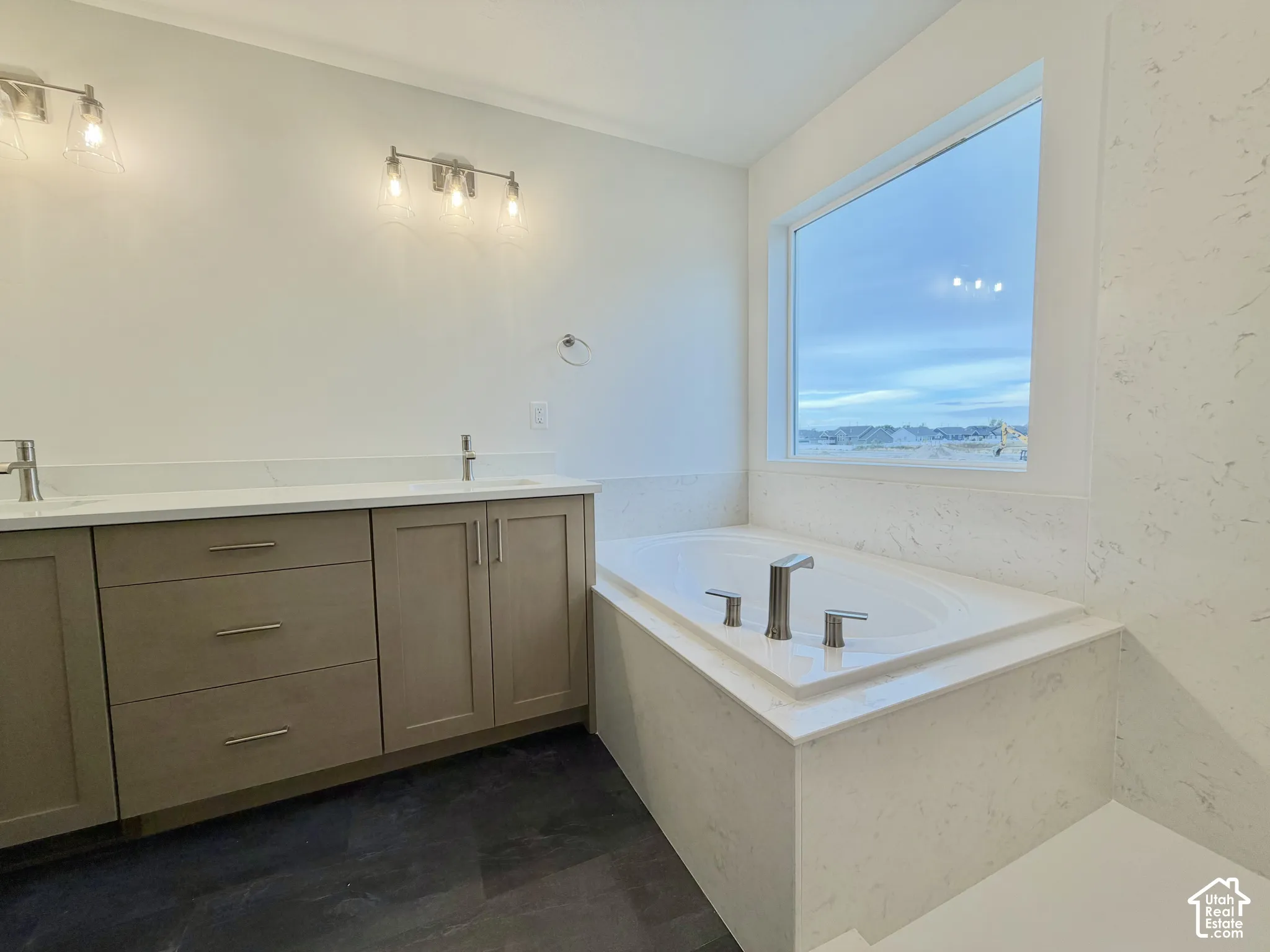 Double vanity with light-tone cabinetry, solid-surface countertops, dual under-mount sinks, built-in soaking tub with stone-finish surround, and wall-mounted vanity lighting
