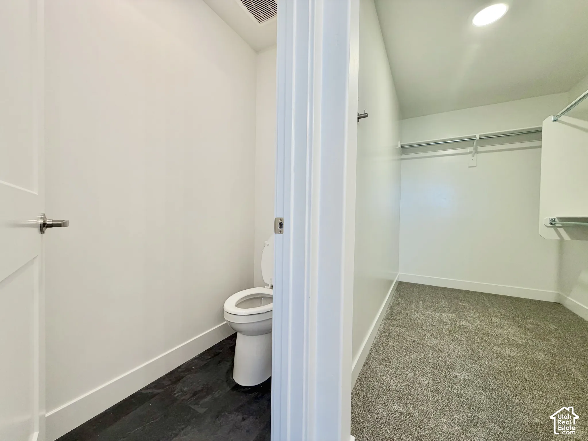 Water closet featuring dark plank-style flooring and a flush toilet, paired with a walk-in closet featuring wall-to-wall carpeting and built-in metal shelving