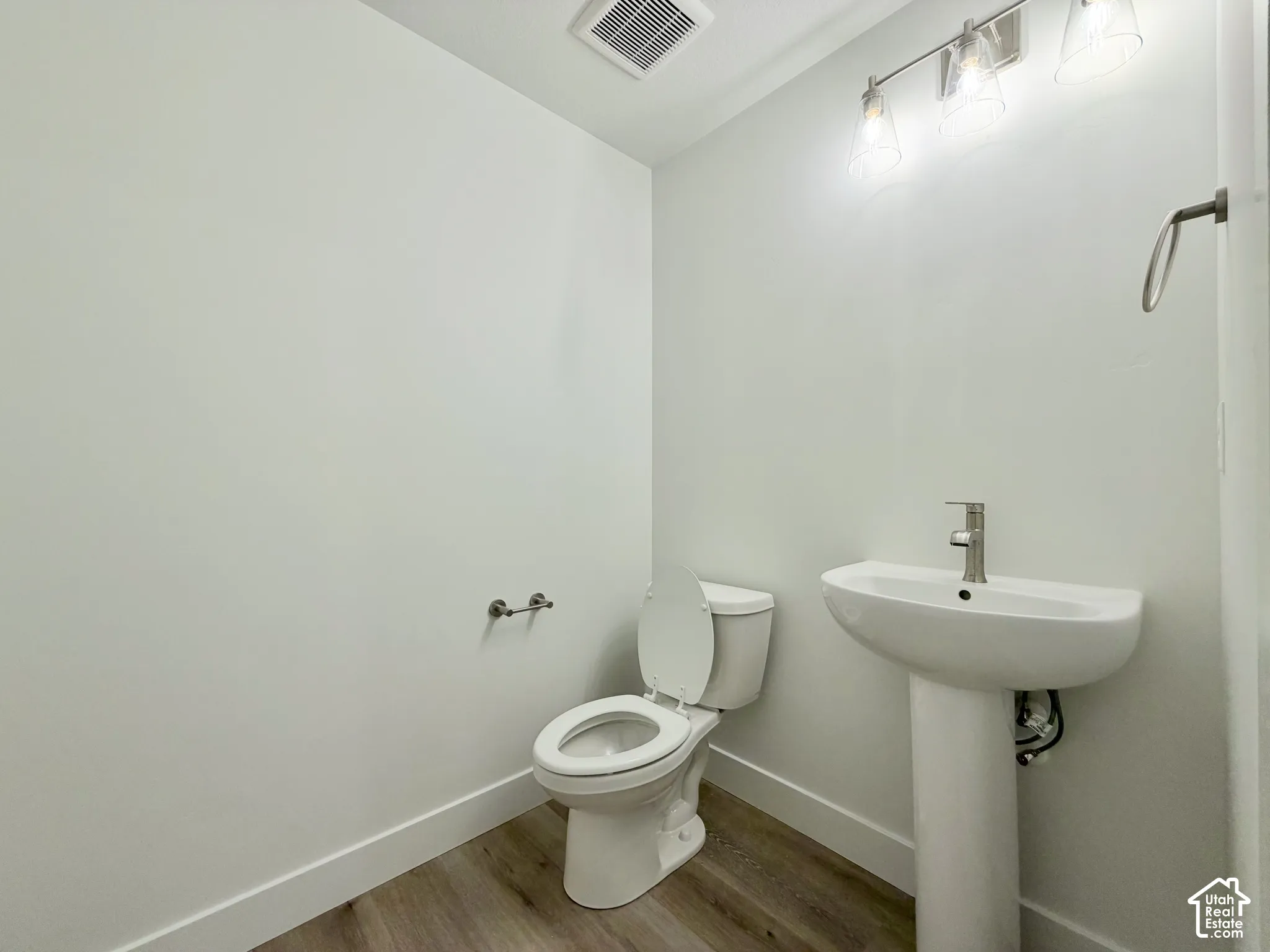 Ceramic pedestal sink with a single-handle chrome faucet, wall-mounted towel ring, white toilet, and wood-finish flooring complemented by a three-light vanity fixture