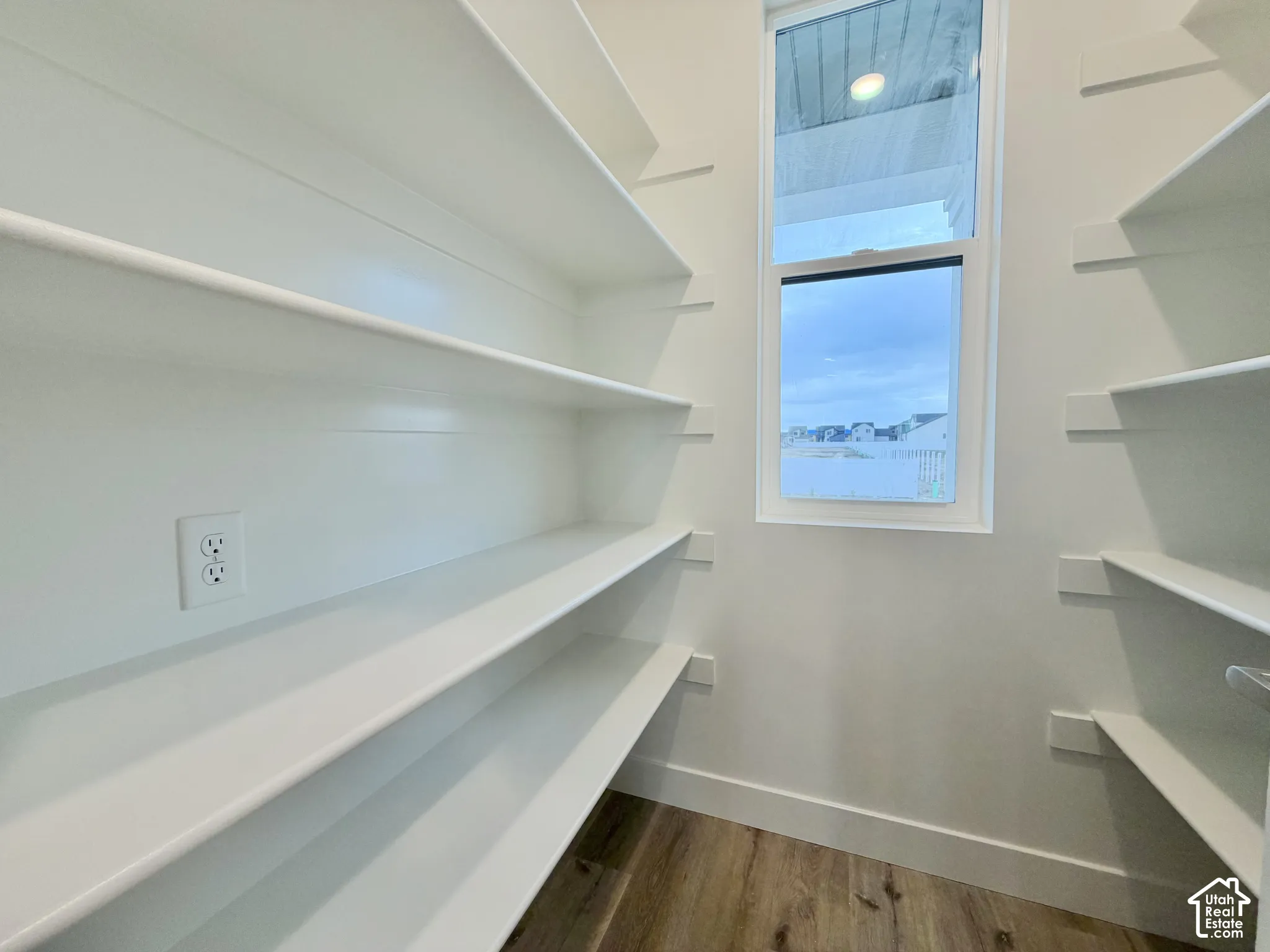 Walk-in storage space featuring multi-level white shelving, wood-finish flooring, and a single-pane window