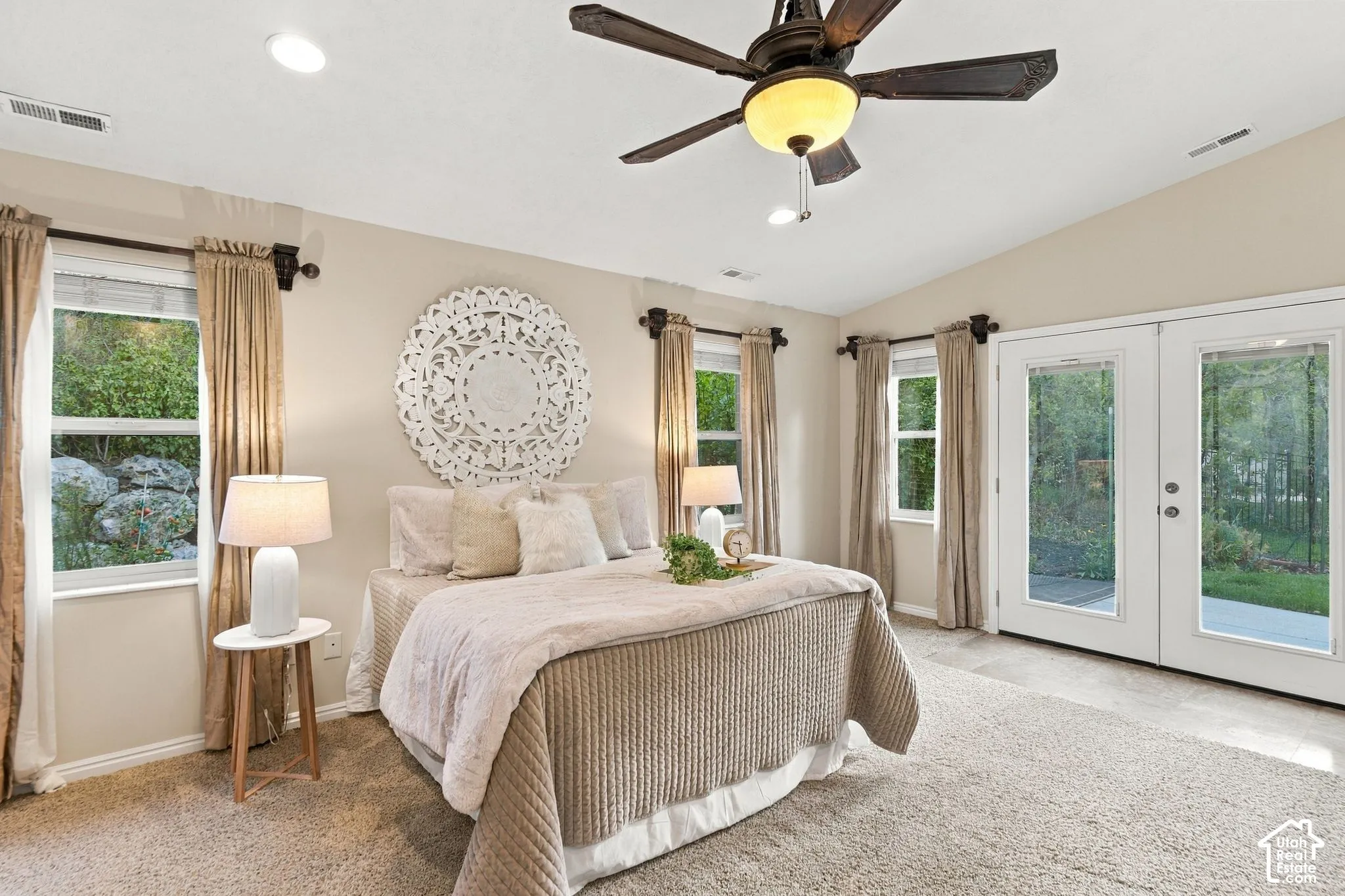 Primary bedroom with vaulted ceilings, neutral wall tones, carpeted flooring, a multi-blade ceiling fan with integrated light fixture, and double glass doors providing outdoor access