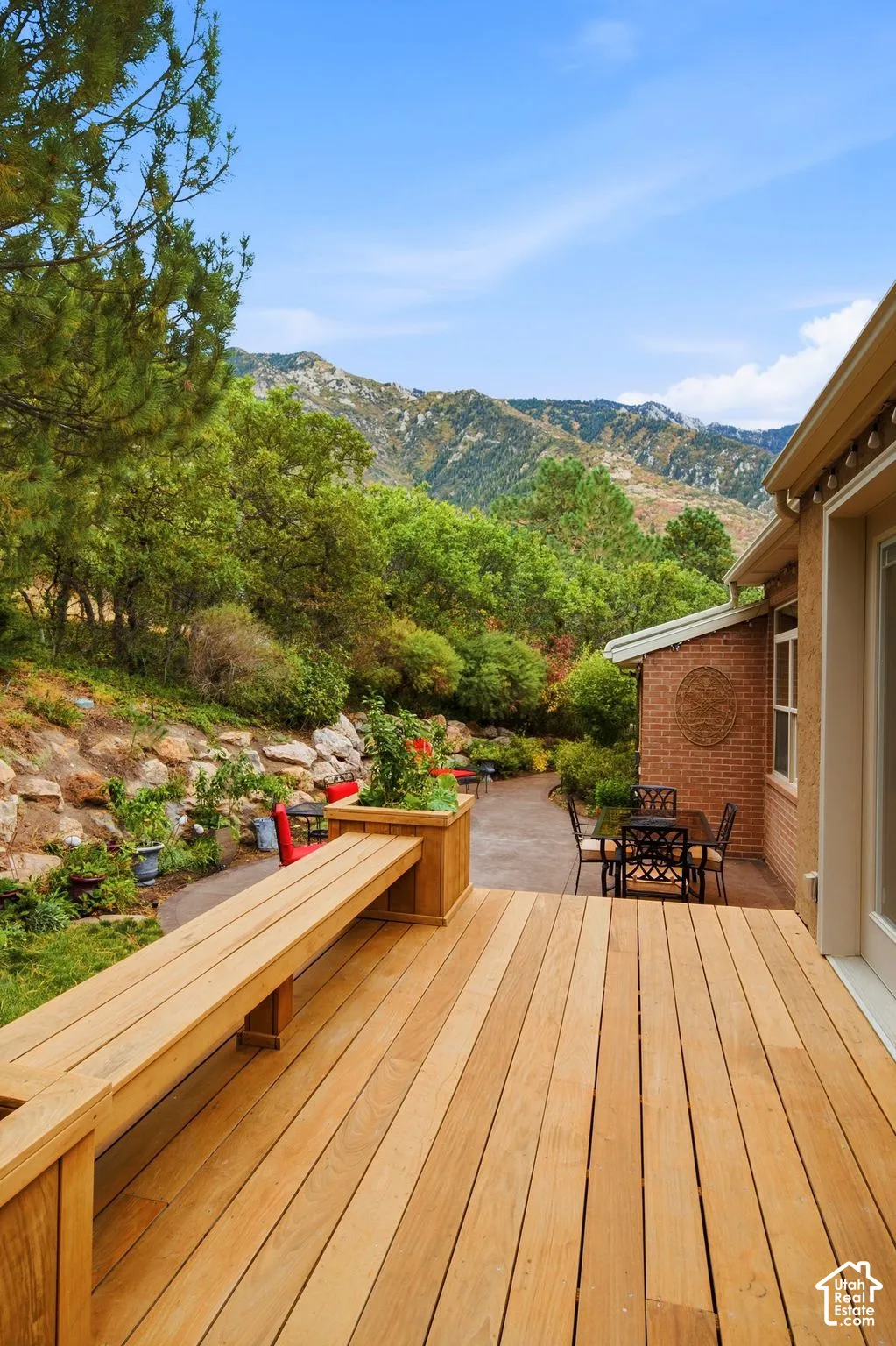 Expansive wood-plank deck featuring a built-in bench, brick exterior wall, stamped concrete patio, and mature trees against a mountain backdrop