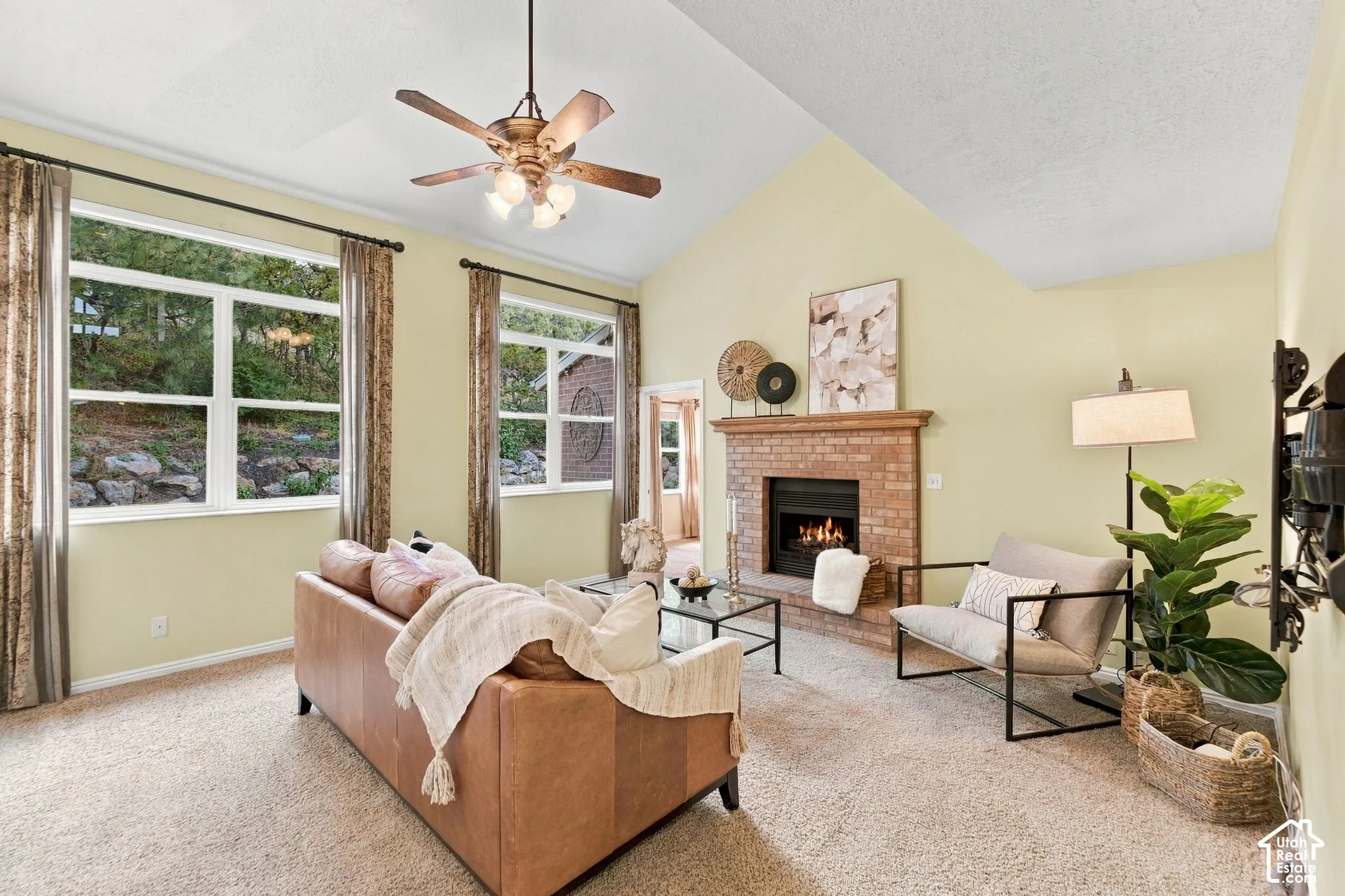 Vaulted ceilings featuring a central ceiling fan, large windows, and a brick fireplace with a wood mantel