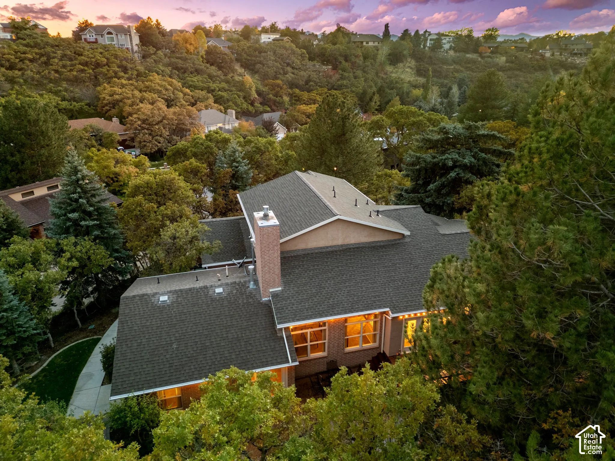 Aerial perspective of a multi-level residence featuring a multi-pitched shingled roof, a brick chimney, and mature surrounding landscaping
