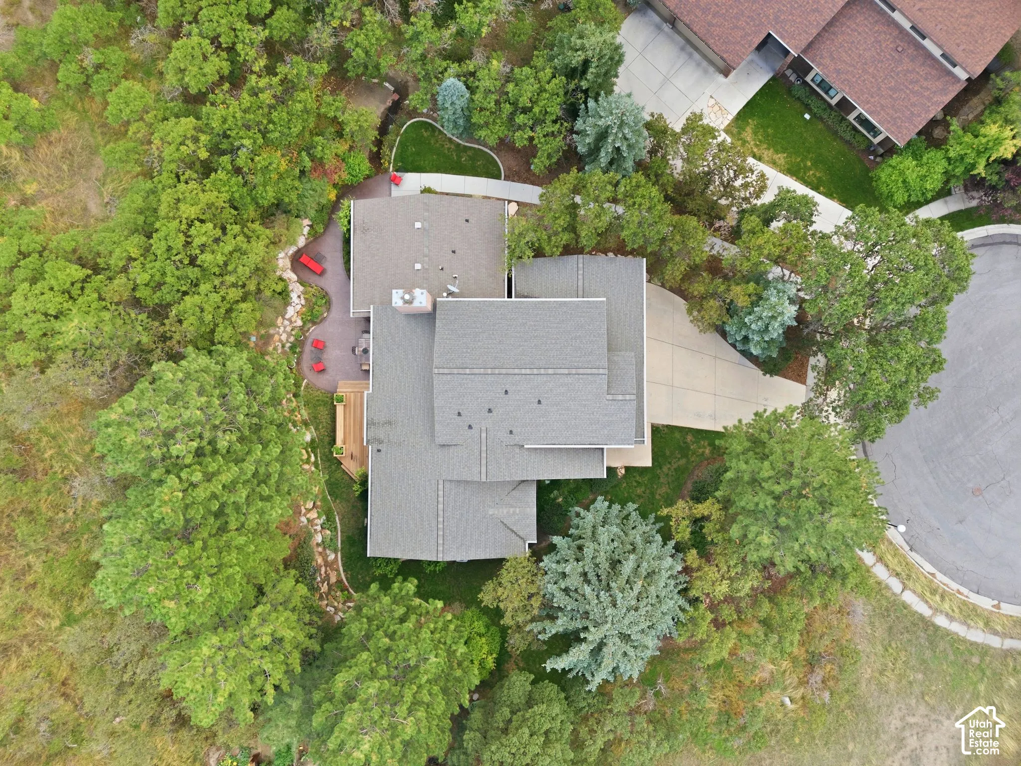 Aerial perspective of a property featuring a multi-level roofline, extensive mature tree cover, a private concrete driveway, hardscaped patio area, and natural stone landscaping elements