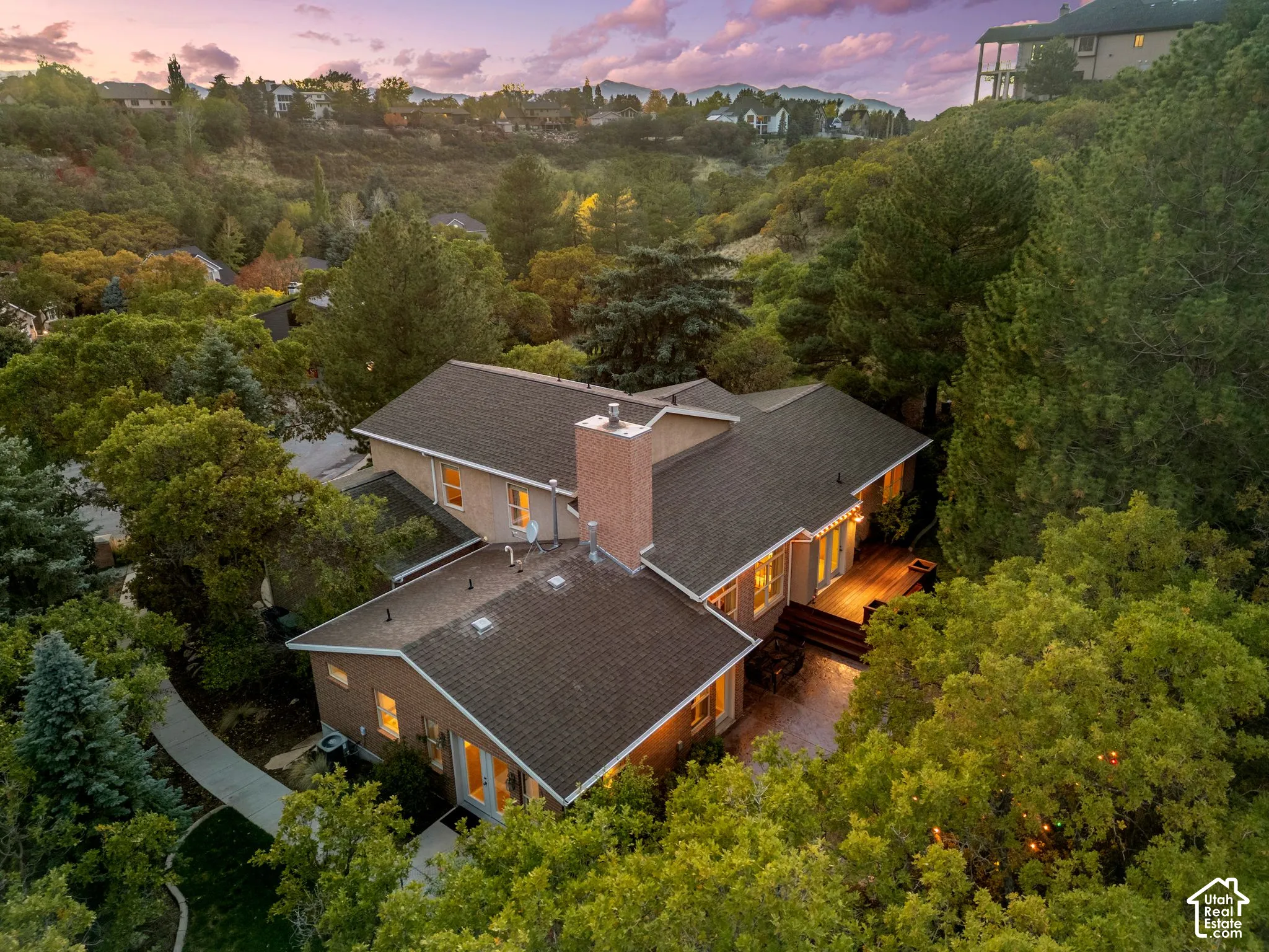 Aerial perspective featuring a multi-level structure with a brick exterior, prominent chimney, shingled roofing, and a wooden deck, set within a landscaped lot with dense mature trees and mountain scenery