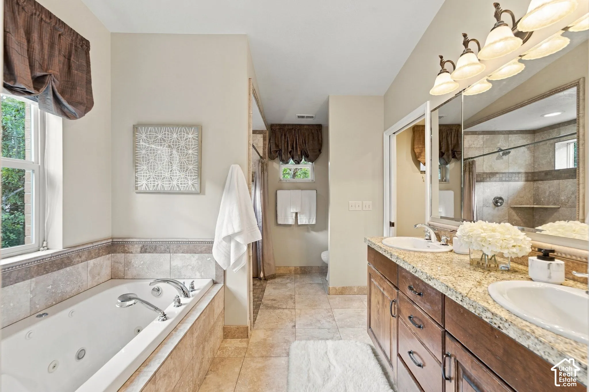 Primary bathroom featuring a jetted soaking tub with tile surround, double-sink vanity with stone countertop, large wall-mounted mirror, and tile flooring