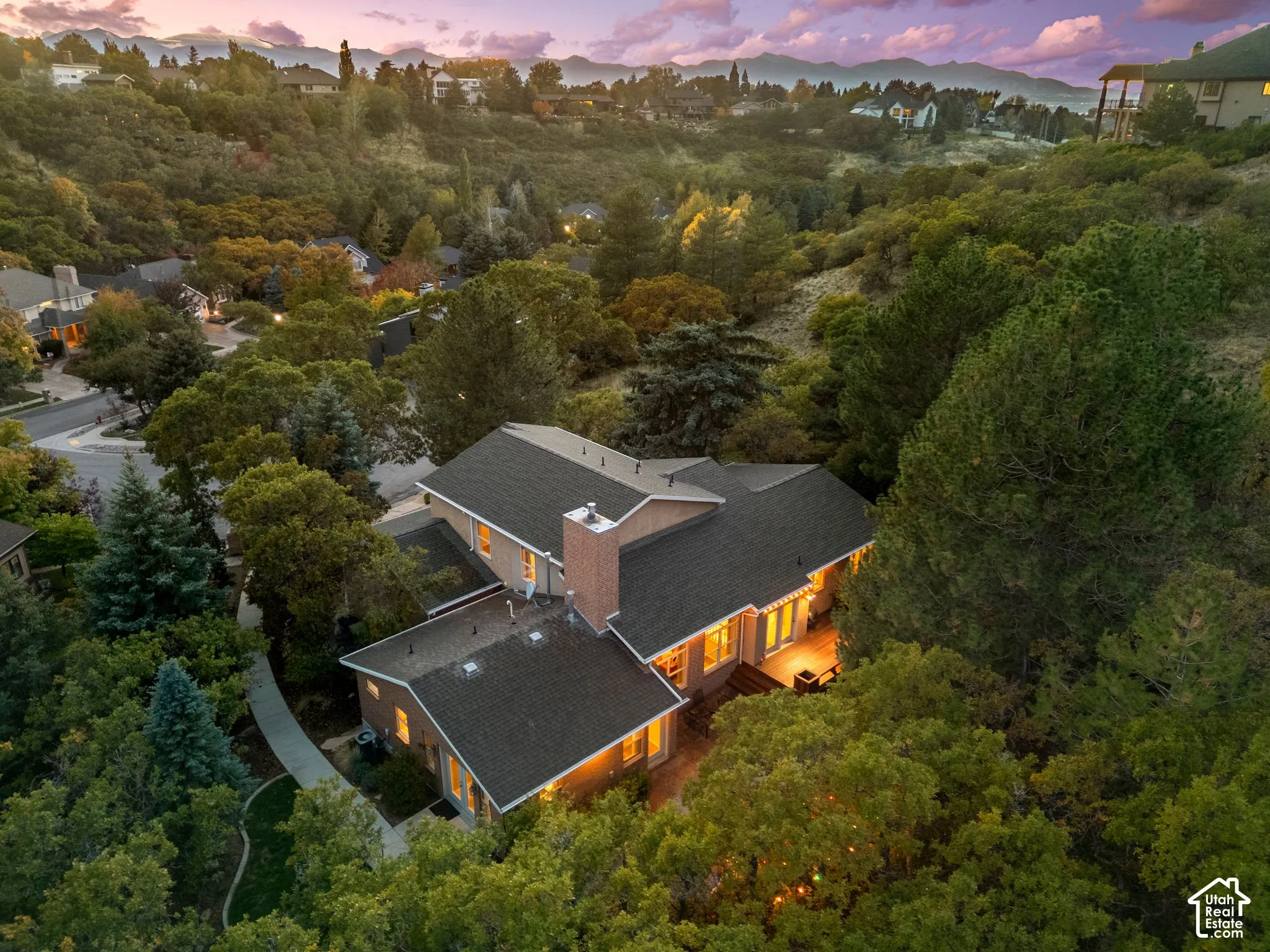 Multi-level residence featuring a brick chimney, exterior wood-finish siding, a rear deck, and mature evergreen and deciduous trees, situated against a mountain backdrop