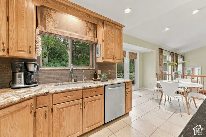 Kitchen featuring natural wood cabinetry, stone countertops, brick-style tile backsplash, stainless steel dishwasher, and ceramic tile flooring