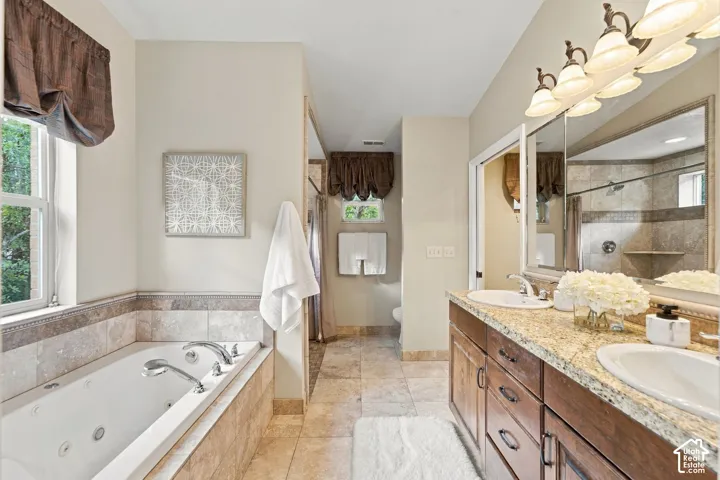 Primary bathroom featuring a jetted soaking tub with tile surround, double-sink vanity with stone countertop, large wall-mounted mirror, and tile flooring
