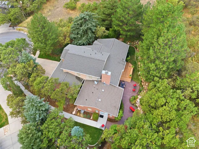 Aerial view of a property featuring a multi-level asphalt shingle roof, exterior brick masonry, a wood-deck platform, a stone paver patio, and mature trees on the surrounding lot