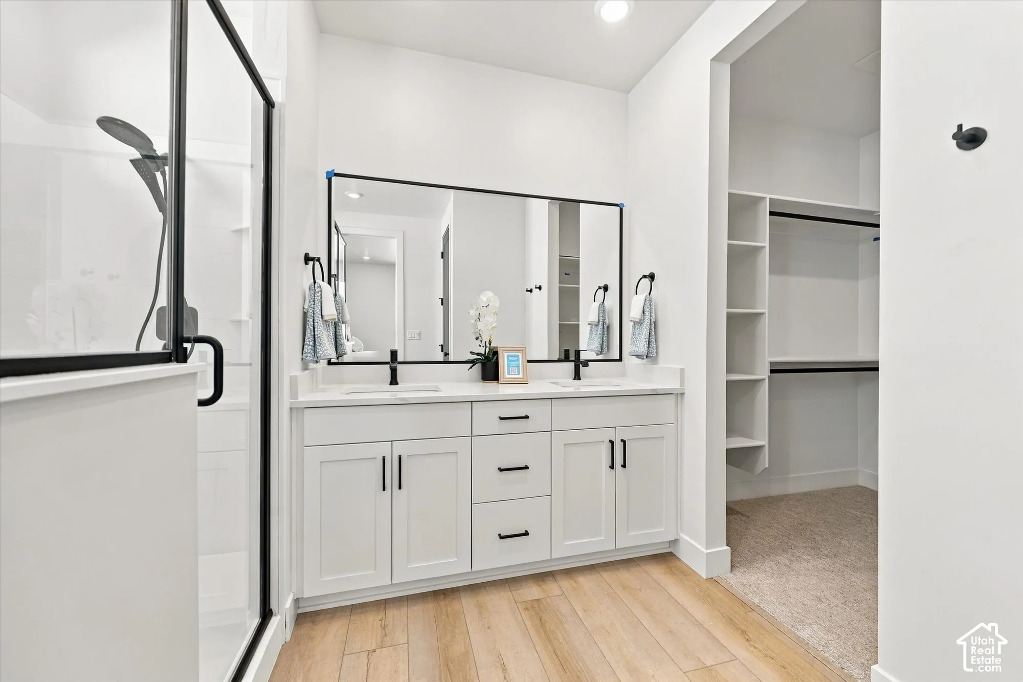 Double vanity with white cabinetry, rectangular vessel sinks, quartz countertops, and matte black hardware, accompanied by a glass-enclosed shower with matte black fixtures, a large-format mirror, and an integrated walk-in closet featuring built-in shelvi