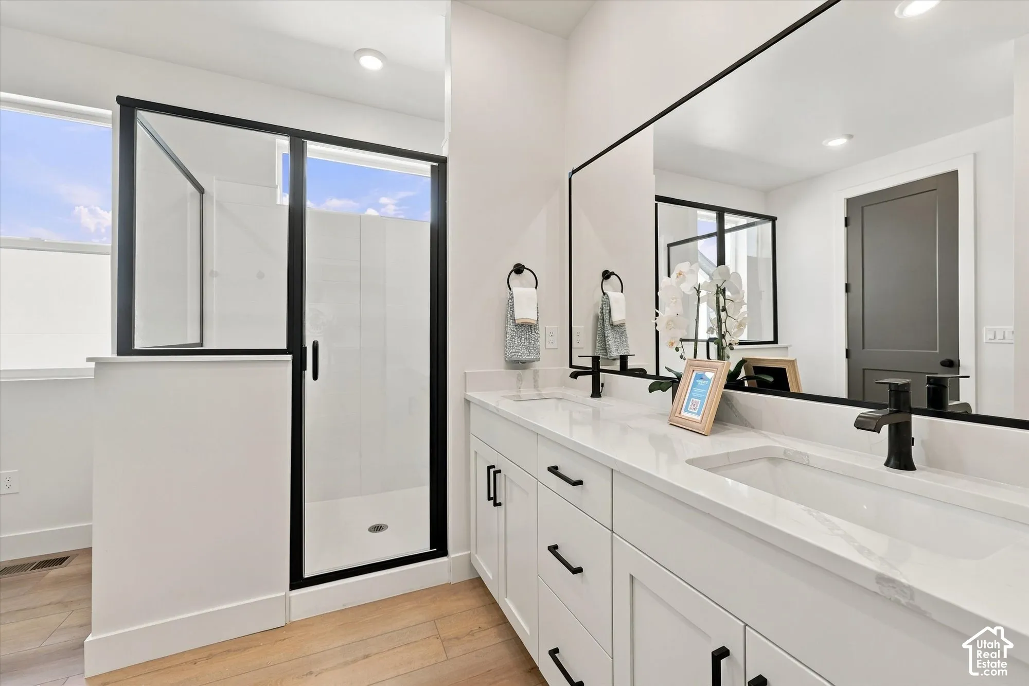 Dual-sink vanity with quartz countertops, white shaker-style cabinetry, matte black hardware, glass-enclosed shower with black frame, and large wall-mounted mirror