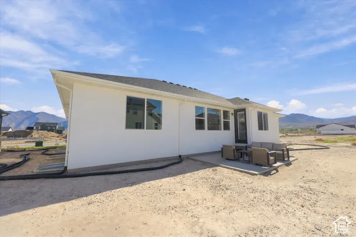Rear exterior featuring white stucco siding, a grey shingle roof, concrete patio, and window-lined facade with mountain scenery