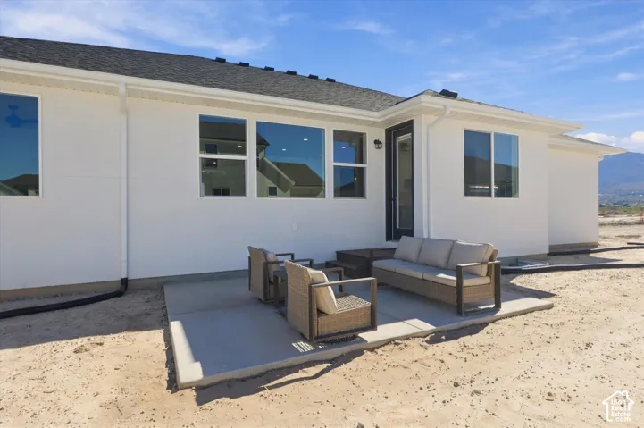 Exterior rear elevation featuring a smooth stucco finish, asphalt shingle roofing, expansive multi-pane windows, and a concrete patio