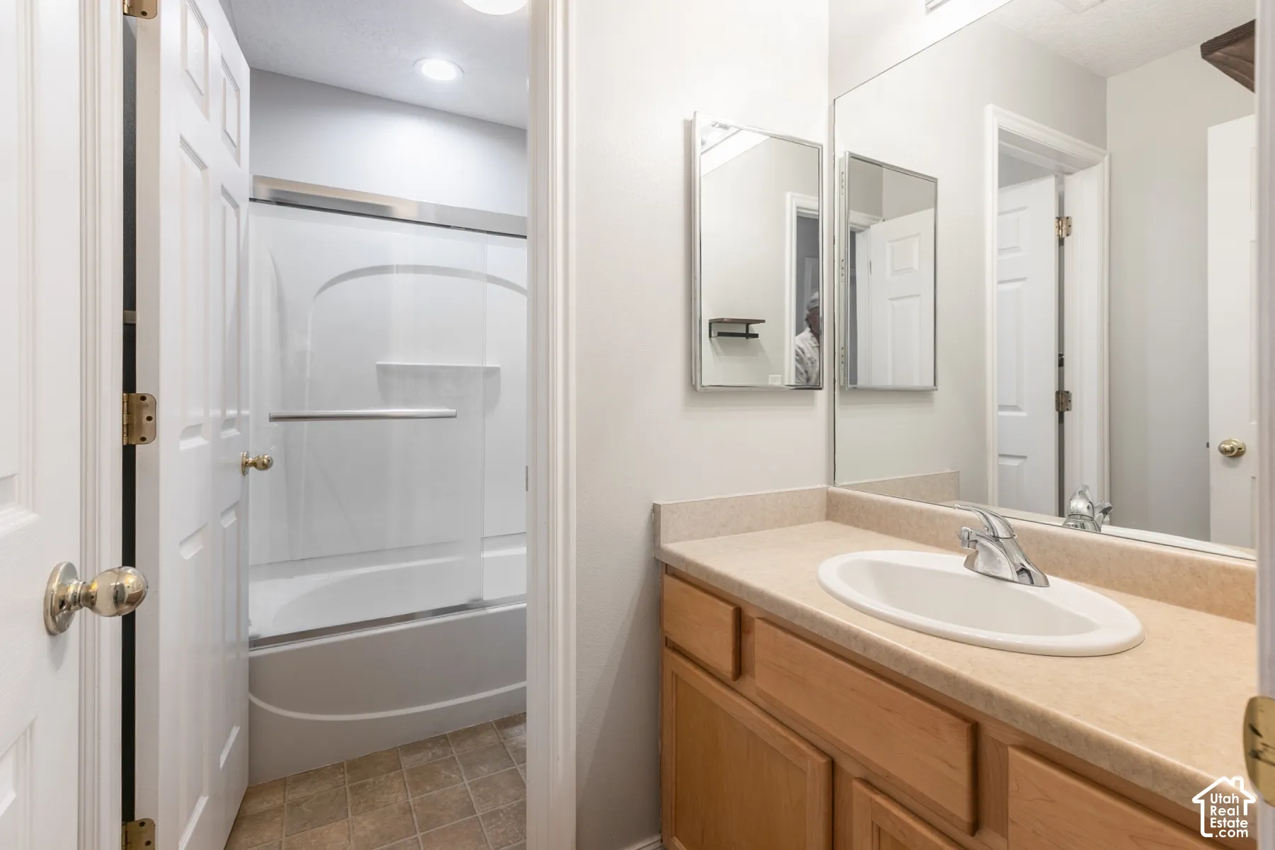Bathroom featuring a vanity with light-tone solid-surface countertop and integrated oval sink, full-width vanity mirror, recessed ceiling lighting, and a tub-shower combination with a sliding glass door