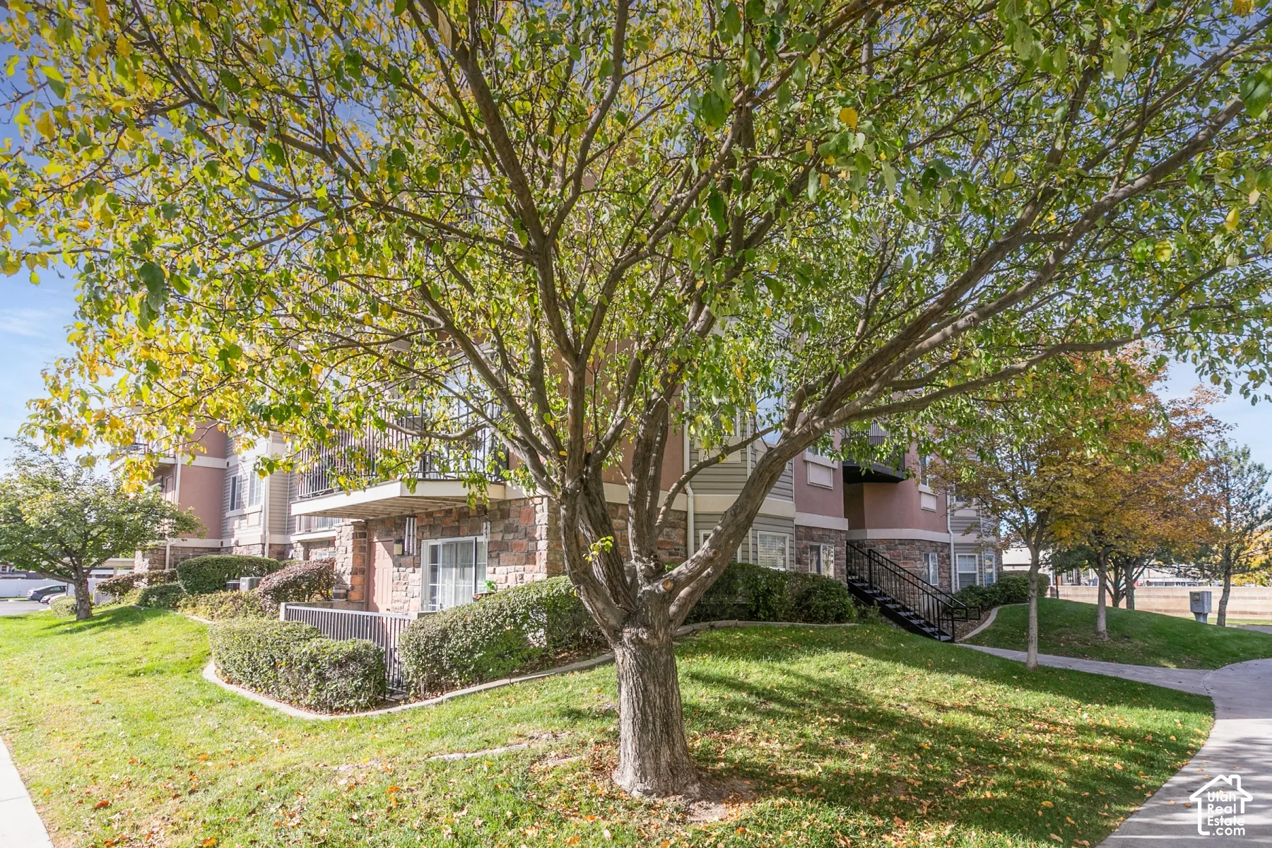 Multi-story exterior featuring stone veneer siding, balconies with wrought-iron railings, manicured landscaping, and mature deciduous trees