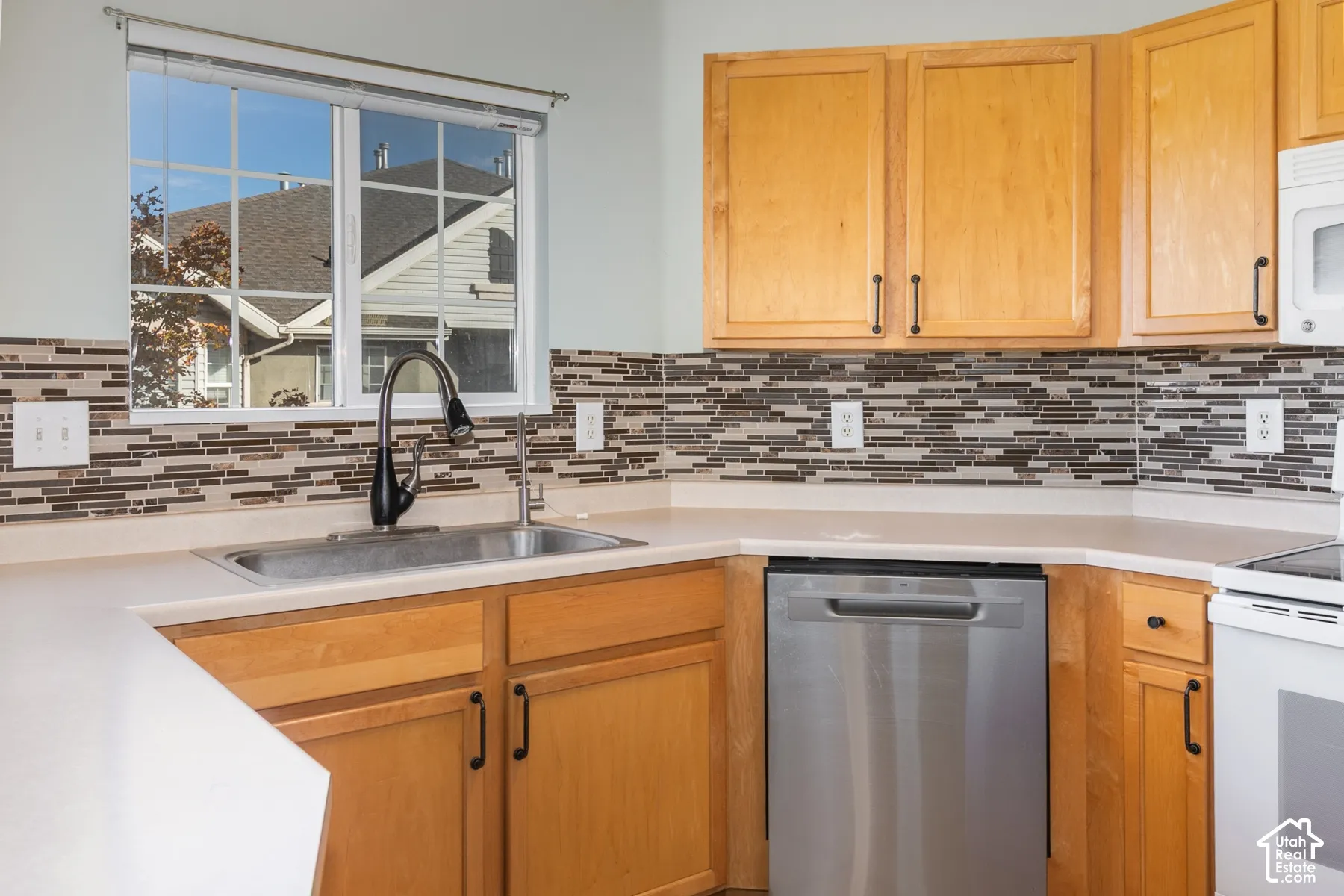 Kitchen featuring shaker-style wood cabinetry, stainless steel dishwasher, linear glass tile backsplash, neutral countertops, and a dual-handle high-arc faucet