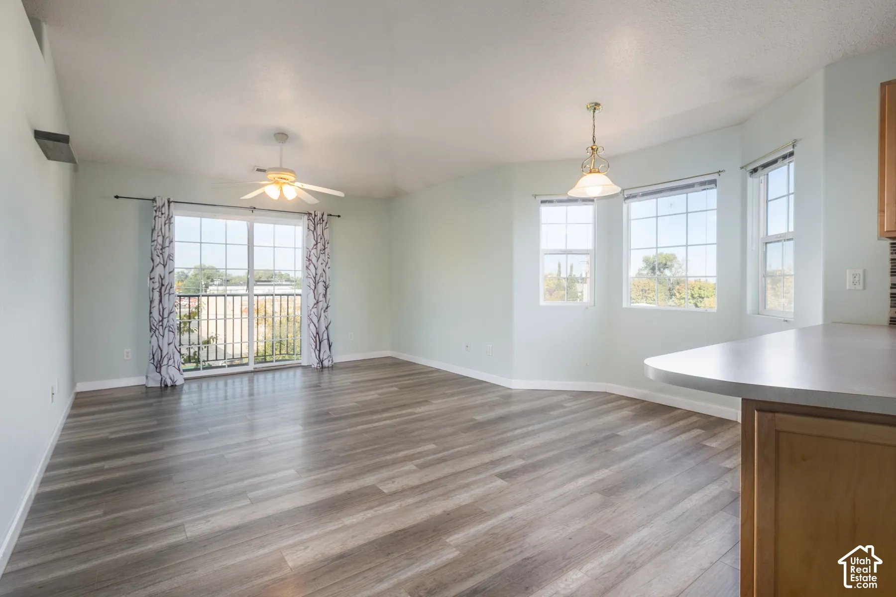 Open interior space featuring wood-finish flooring, sliding glass patio door, bay-style window configuration, ceiling fan with integrated light, and overhead pendant lighting