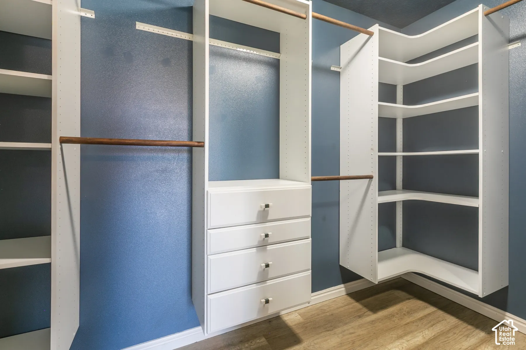 Custom closet featuring white built-in shelving, a four-drawer storage unit, wood-finish closet rods, and wood-finish flooring