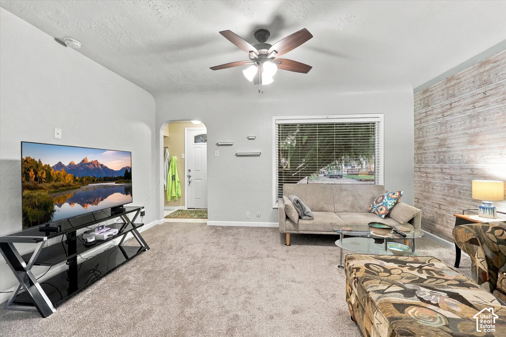 Living space featuring a natural-finish wood plank accent wall, neutral carpeting, a central ceiling fan with integrated lighting, and an arched entryway