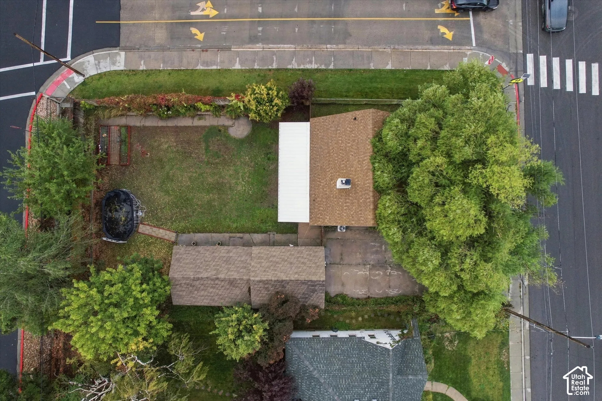 Aerial view of a corner lot property featuring mature trees, an expansive lawn, a paved driveway, and a shingle-roofed structure