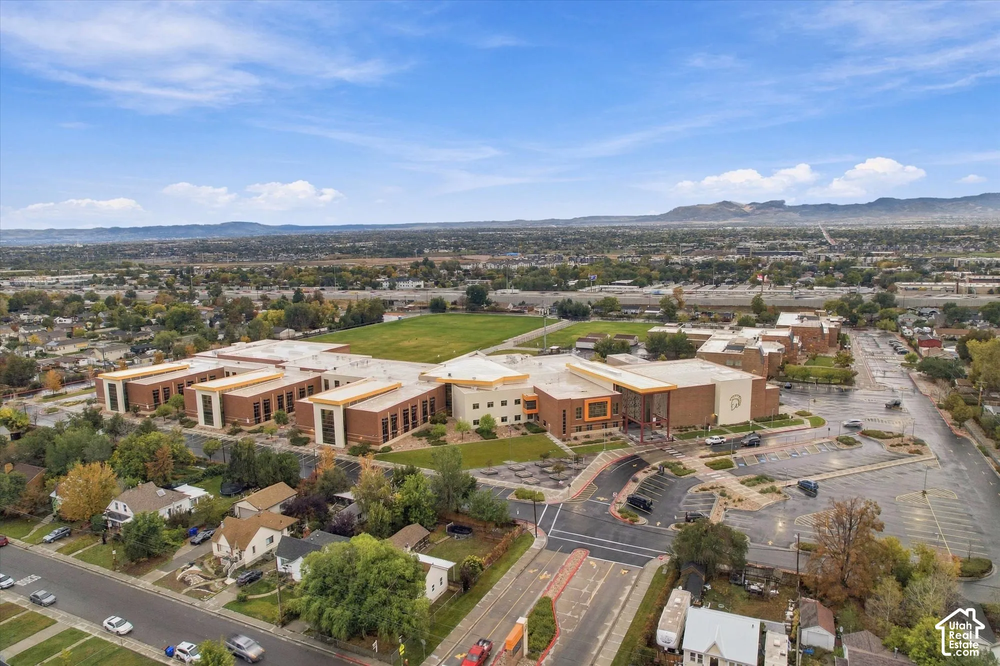 Aerial perspective of the large brick-facade facility featuring extensive paved parking surfaces, expansive open green space, multi-sectioned flat roofing, and a prominent mountain backdrop