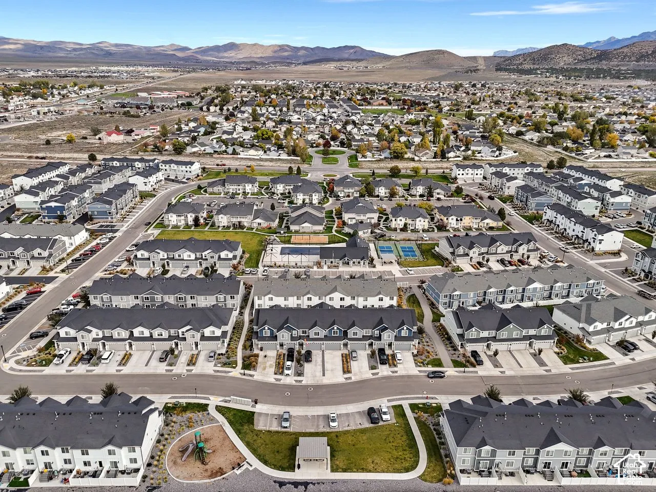 Aerial perspective of a multi-unit residential community featuring a central swimming pool, hard-surfaced sports courts, a playground, and mountain range scenery