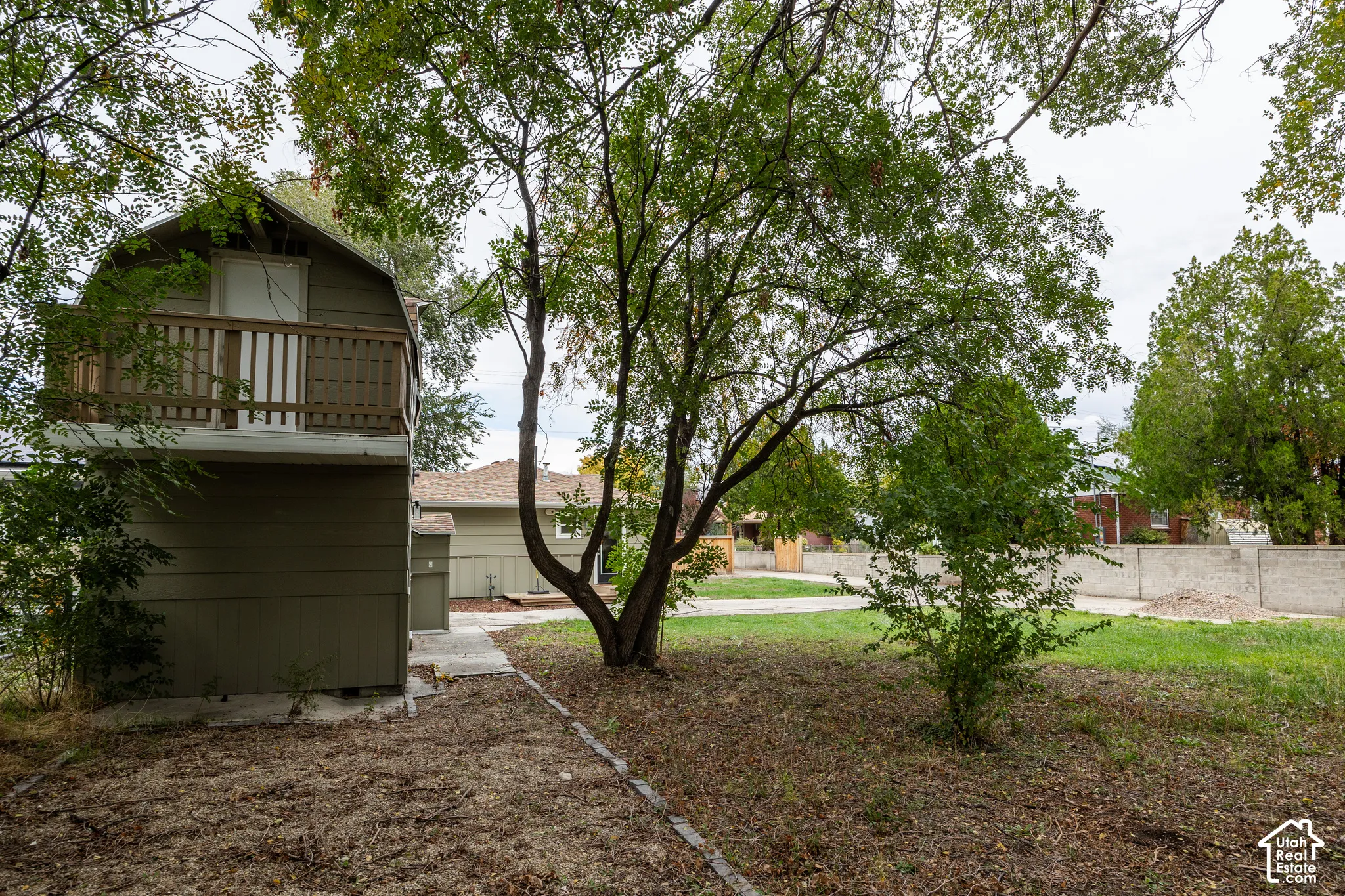Detached structure featuring a second-story wooden balcony, concrete siding, and a gabled roof, situated on a lot with mature trees and a masonry perimeter wall