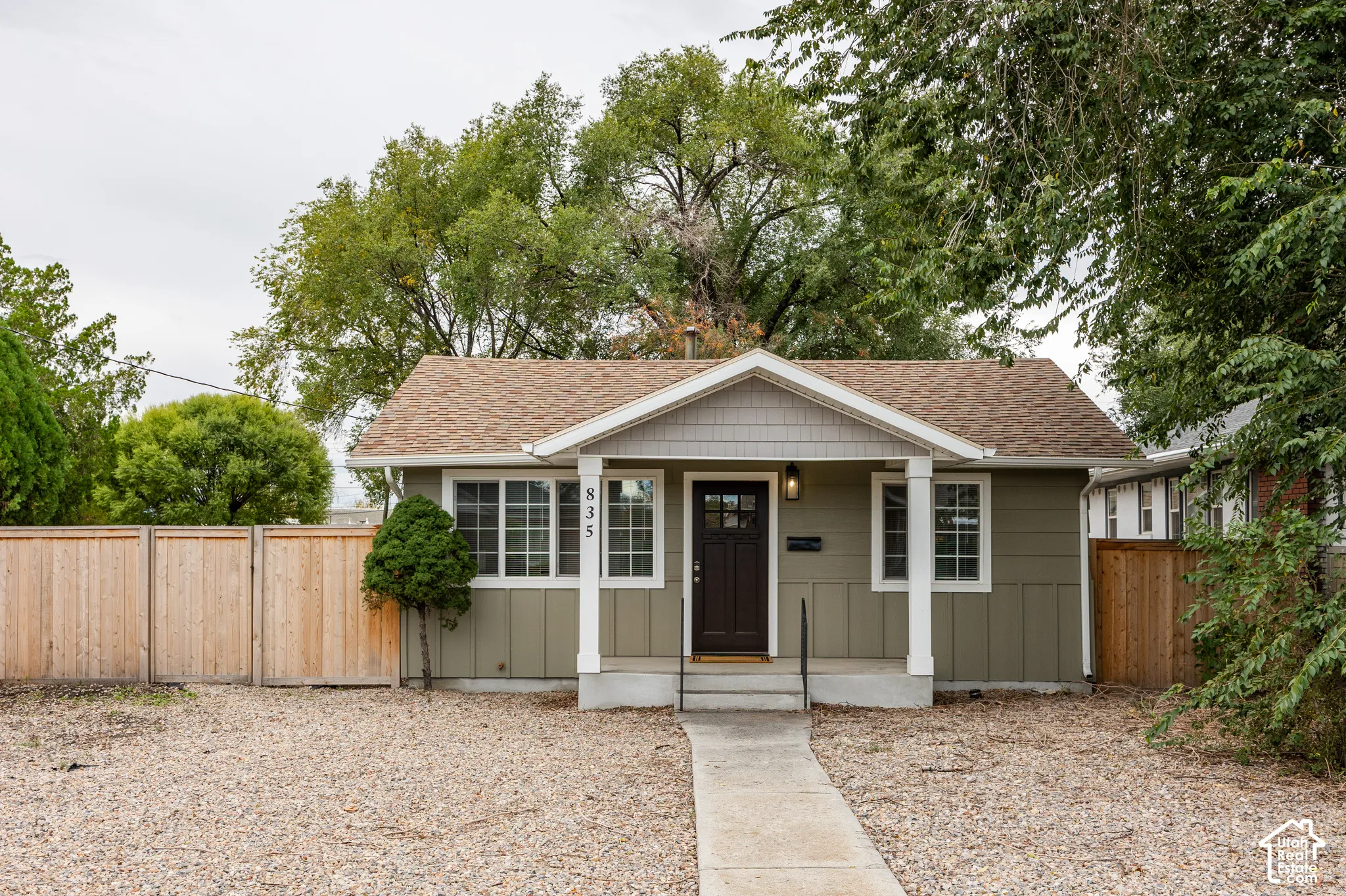 Single-story exterior featuring composite siding, shingle roofing, a covered front porch with square columns, and wood-finish privacy fencing