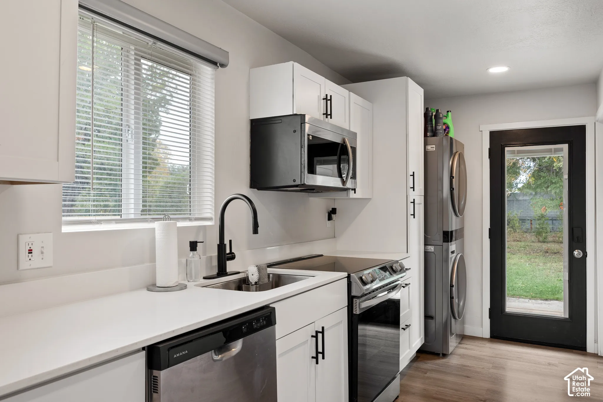 Laundry area featuring white shaker-style cabinetry, solid-surface countertops, a stainless steel undermount sink with a matte black faucet, and wood-finish flooring