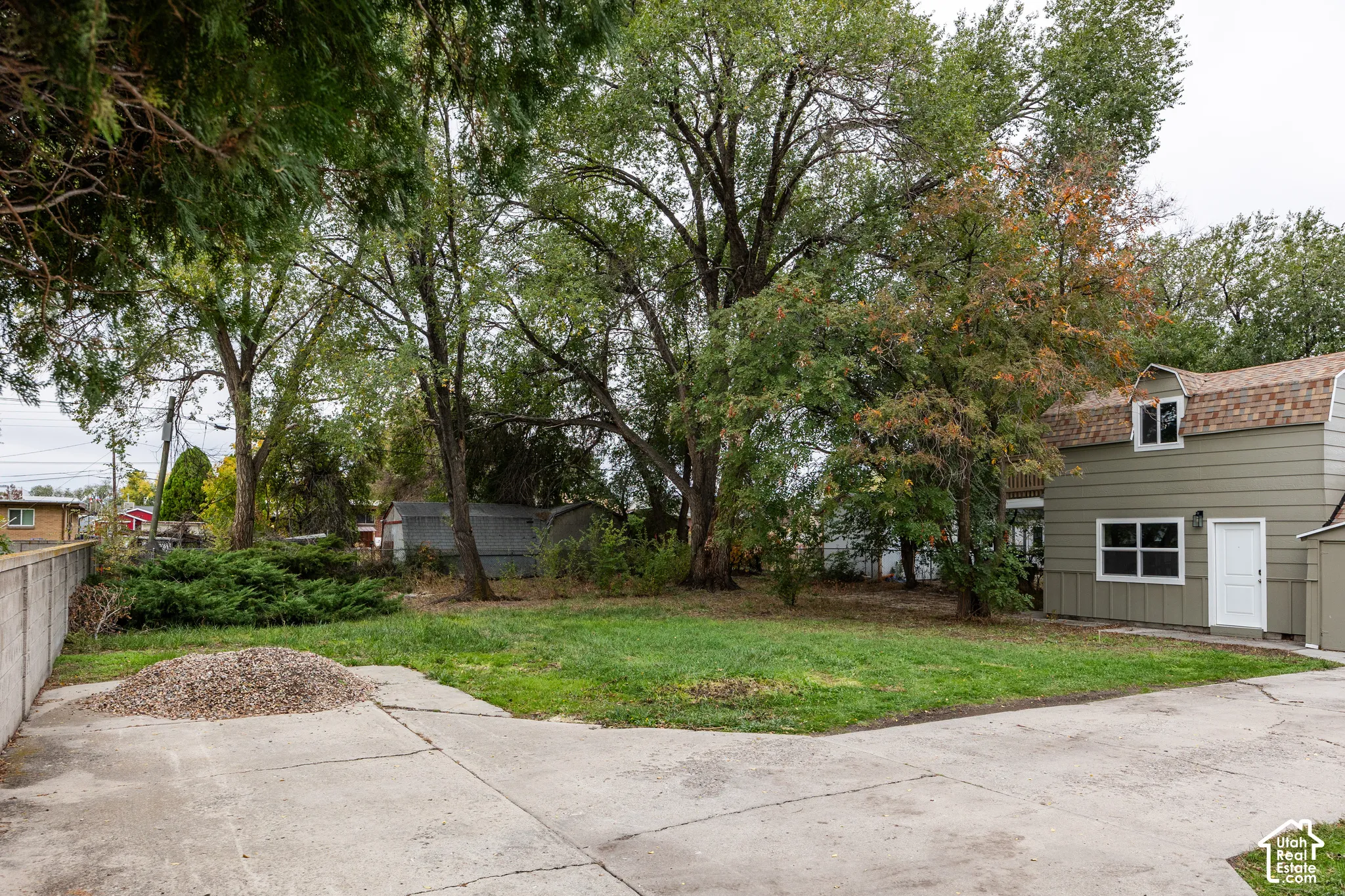Backyard featuring a concrete parking area, mature trees, green lawn, and detached two-story building with shingle roof