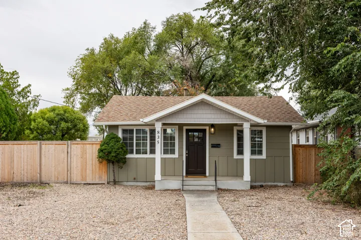 Single-story exterior featuring composite siding, shingle roofing, a covered front porch with square columns, and wood-finish privacy fencing