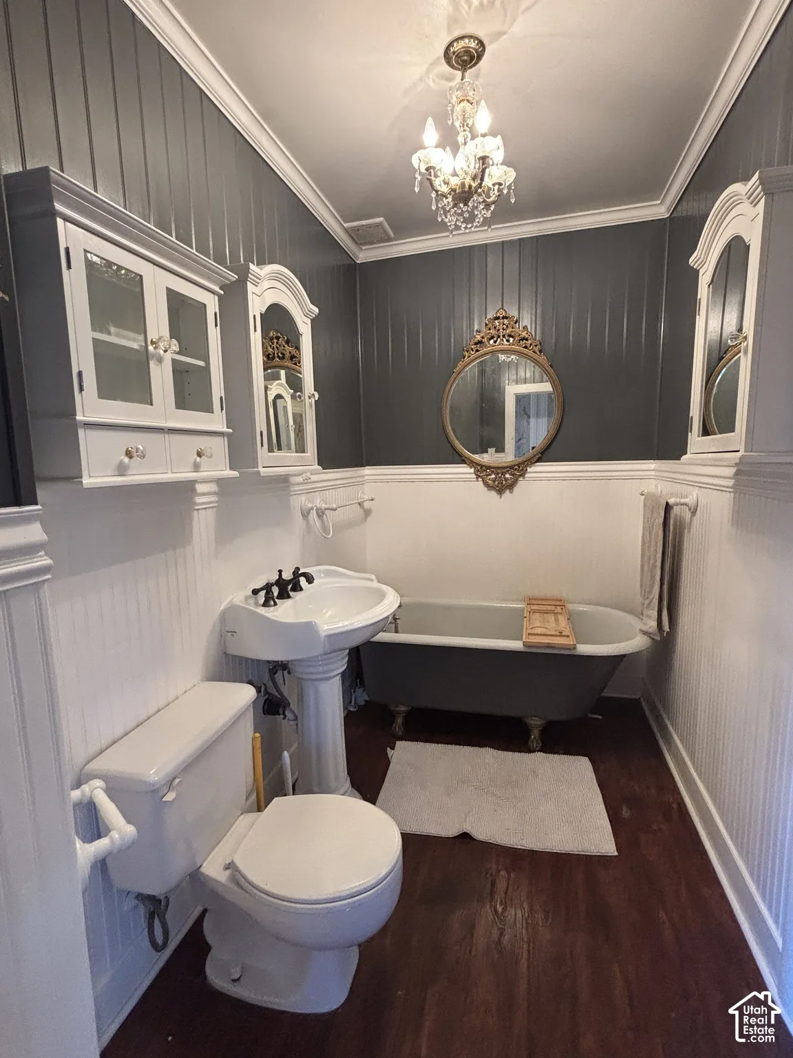 Clawfoot soaking tub, pedestal sink, beadboard wainscoting, ornate wall mirror, and crystal chandelier