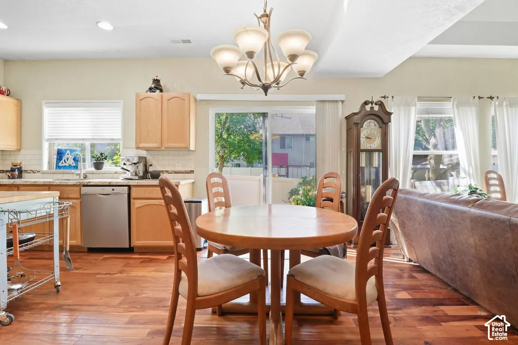 Wood-finish flooring, light wood cabinetry, white subway tile backsplash, stainless steel dishwasher, and a multi-arm chandelier