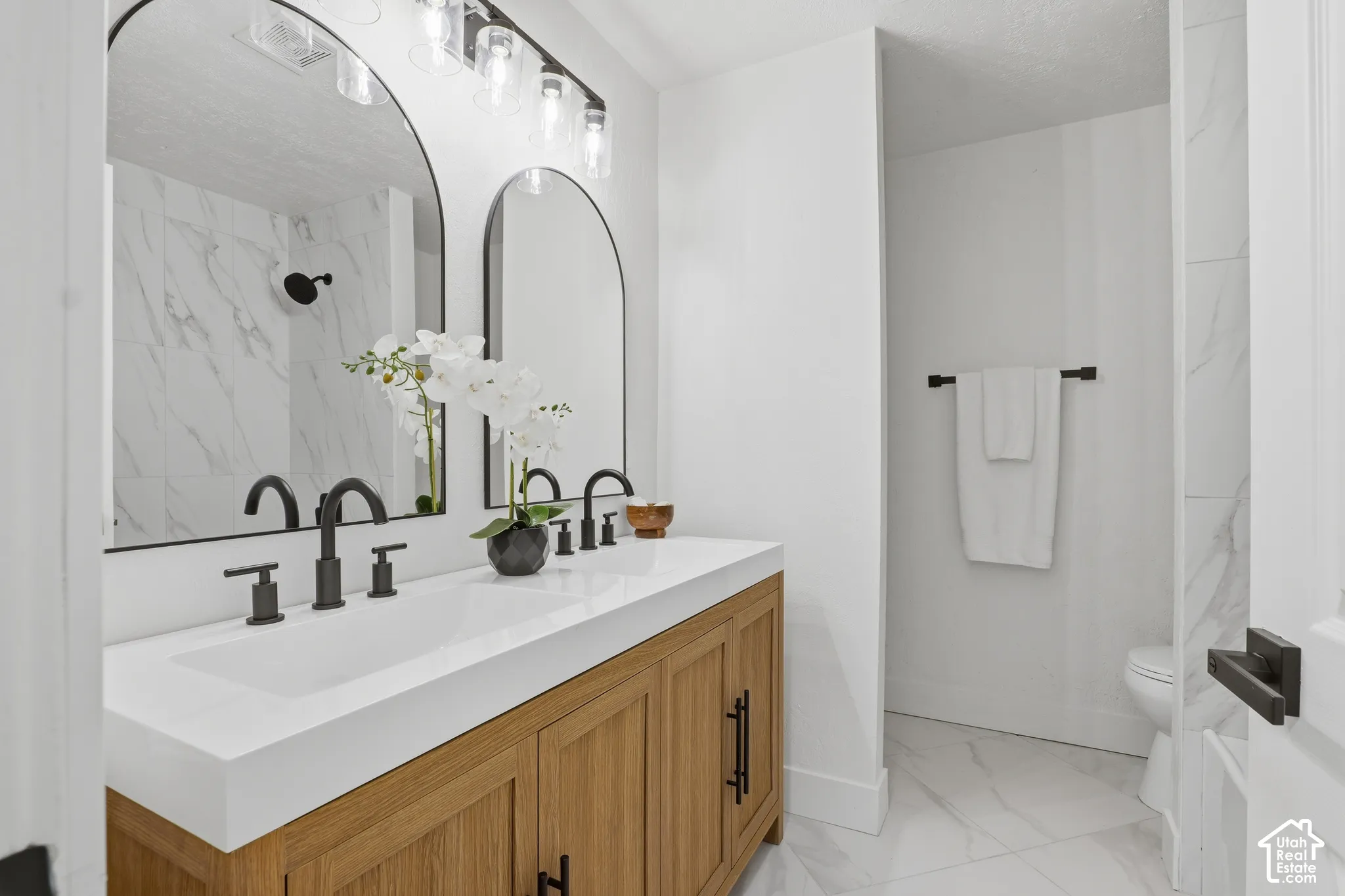 Dual-sink vanity with matte black hardware, arched vanity mirrors, stone-composite countertop, light-toned wood cabinetry, and large-format tile flooring
