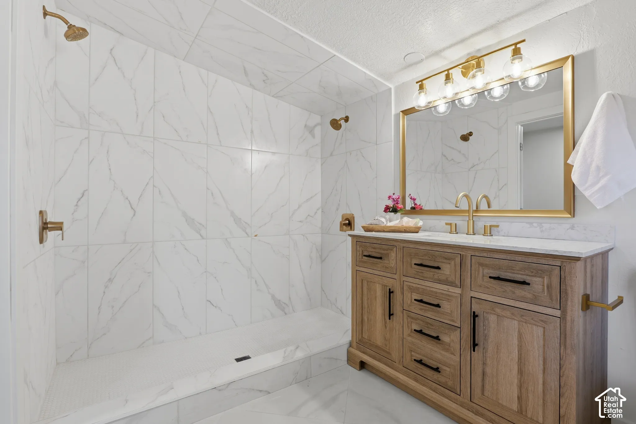 Walk-in shower with floor-to-ceiling large-format tile, wood-finish vanity with brass-tone hardware, quartz-surface countertop, and gold-finish plumbing fixtures