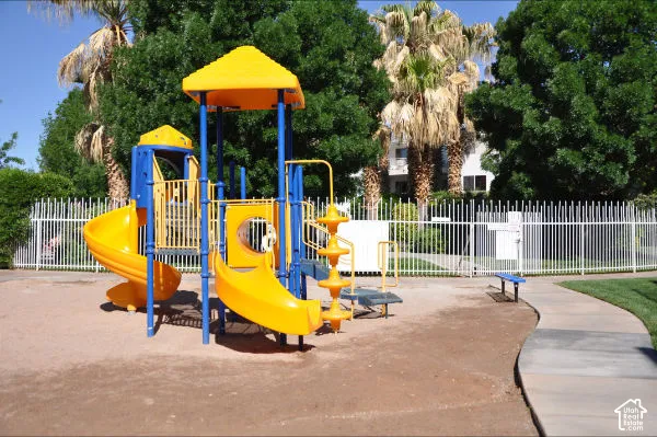 Playground area featuring a multi-level structure with slides, a surrounding white picket fence, a concrete pathway, and a blue park bench
