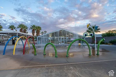 Concrete splash pad with multiple water features, including colorful arches and a frog-themed sprayer, set against a backdrop of palm trees and a large glass-fronted building