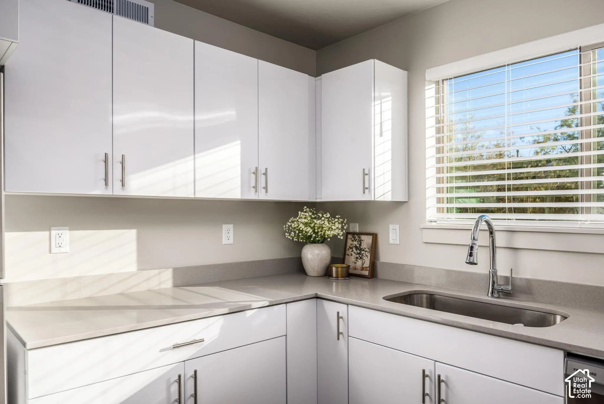 High-gloss white cabinetry featuring brushed metal hardware, solid-surface countertops, undermount stainless steel sink, pull-down chrome faucet, and window with adjustable blinds