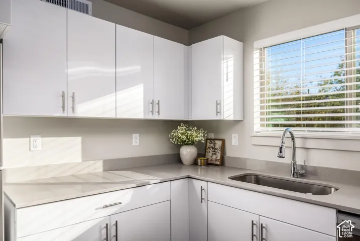 High-gloss white cabinetry featuring brushed metal hardware, solid-surface countertops, undermount stainless steel sink, pull-down chrome faucet, and window with adjustable blinds