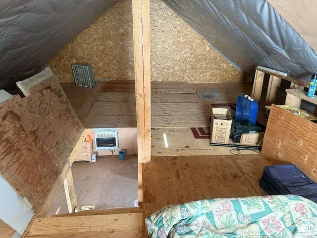Attic space featuring plywood flooring, exposed timber support beams, and wall-mounted heating unit