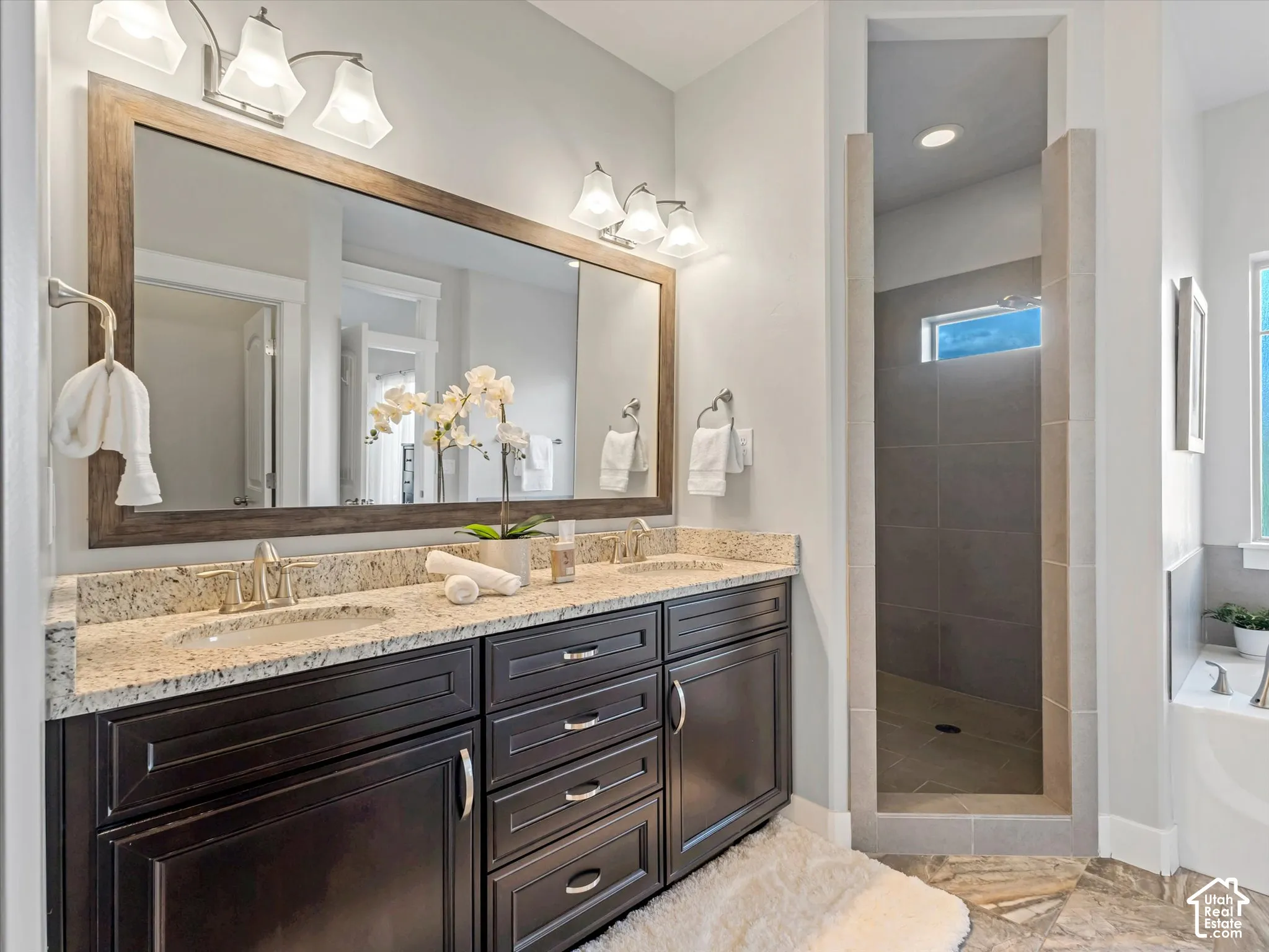 Double vanity featuring dark-toned cabinetry, light-toned stone countertops, dual under-mount sinks, and a full-width framed mirror with overhead vanity lighting