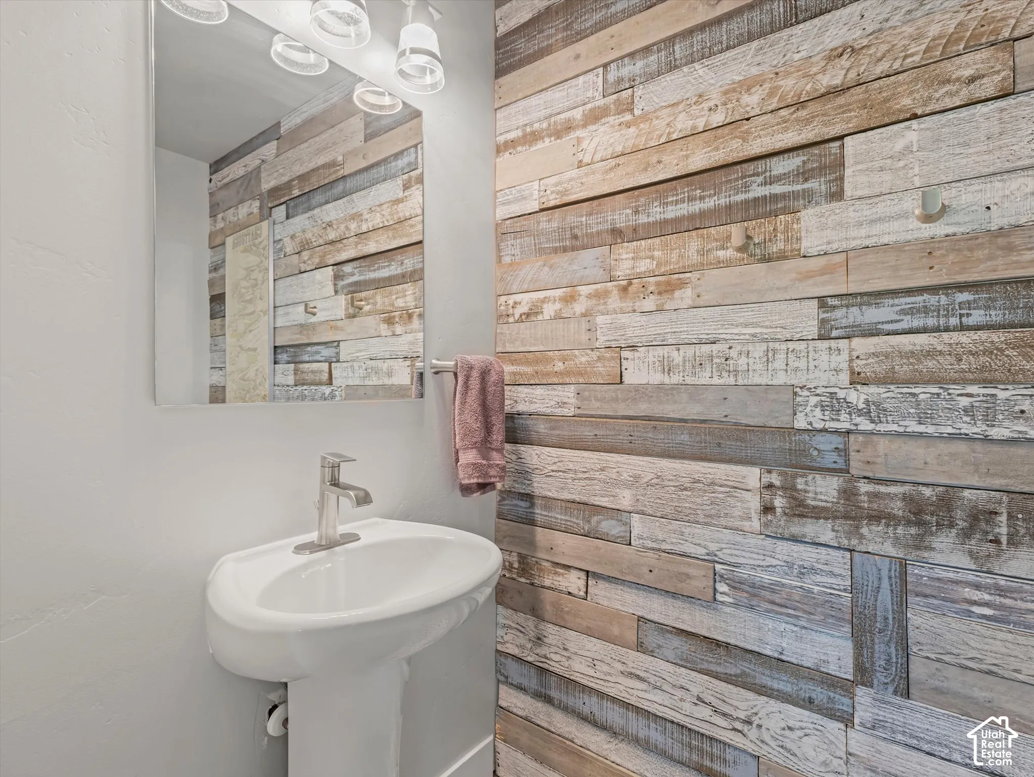 Accent wall featuring horizontal reclaimed wood planks, white pedestal sink, polished chrome faucet, wall-mounted mirror, and multi-bulb light fixture