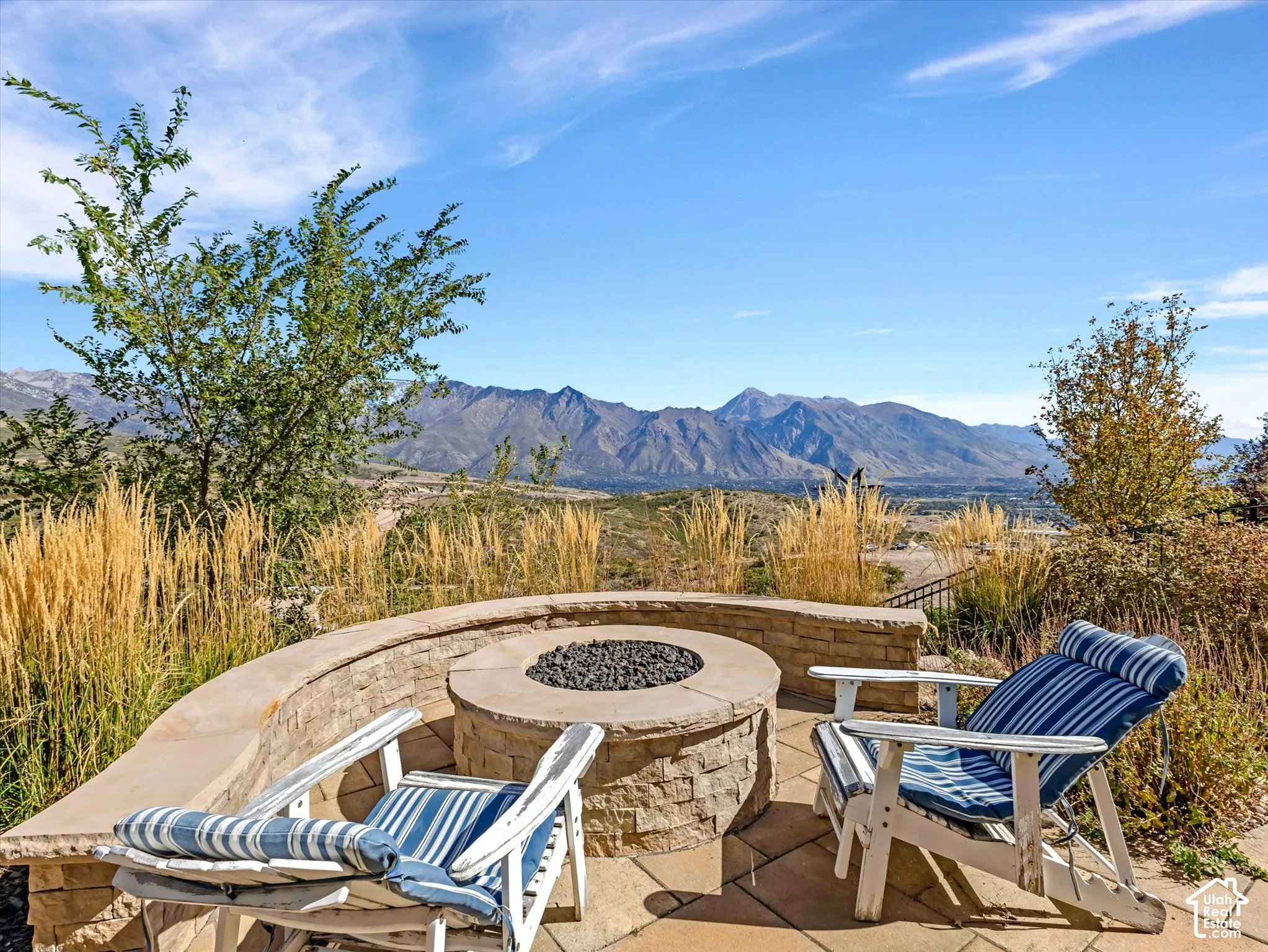 Circular stone fire pit featuring a stacked-stone base and cap, set within a curved stone retaining wall on a paved patio, complemented by mountain scenery