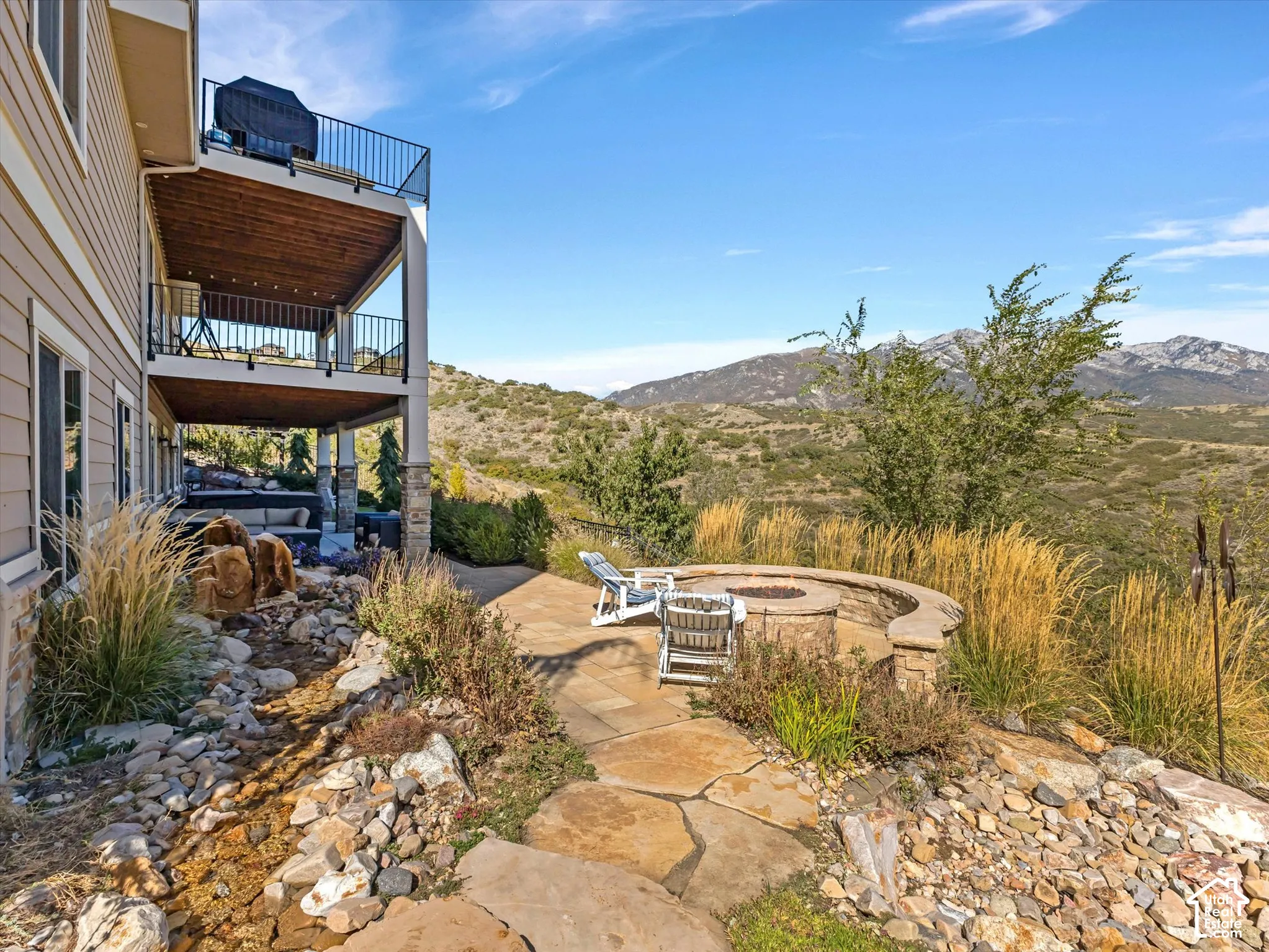 Multi-level rear balcony with metal railings and wood-paneled ceilings, stone-paved patio featuring a built-in circular fire pit with seating wall, and decorative rock landscape elements set against a mountain backdrop