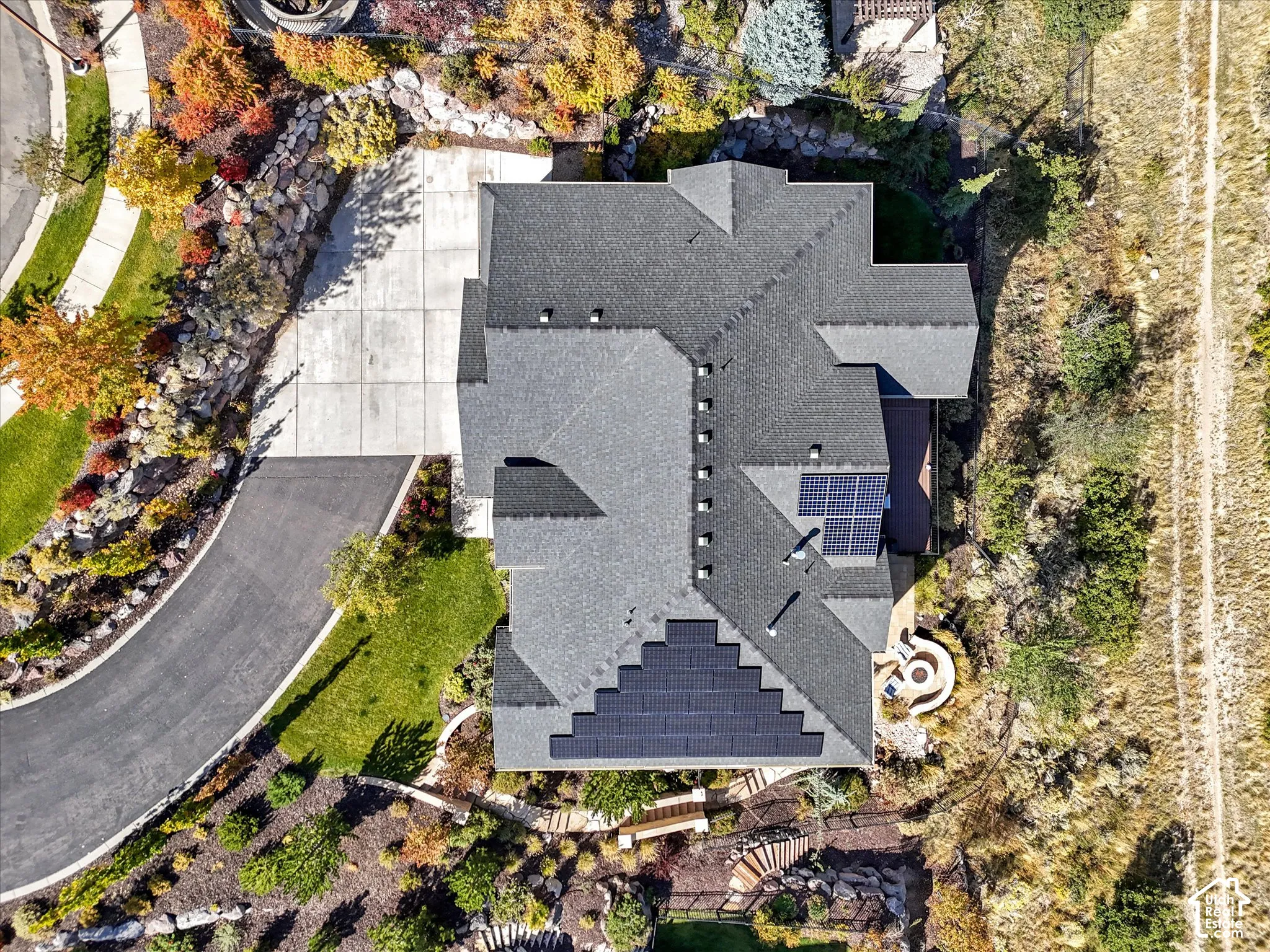 Aerial perspective featuring a multi-faceted shingle roof, integrated solar panel arrays, a multi-car concrete driveway, a paved walkway, and a tiered rock-retaining wall landscape