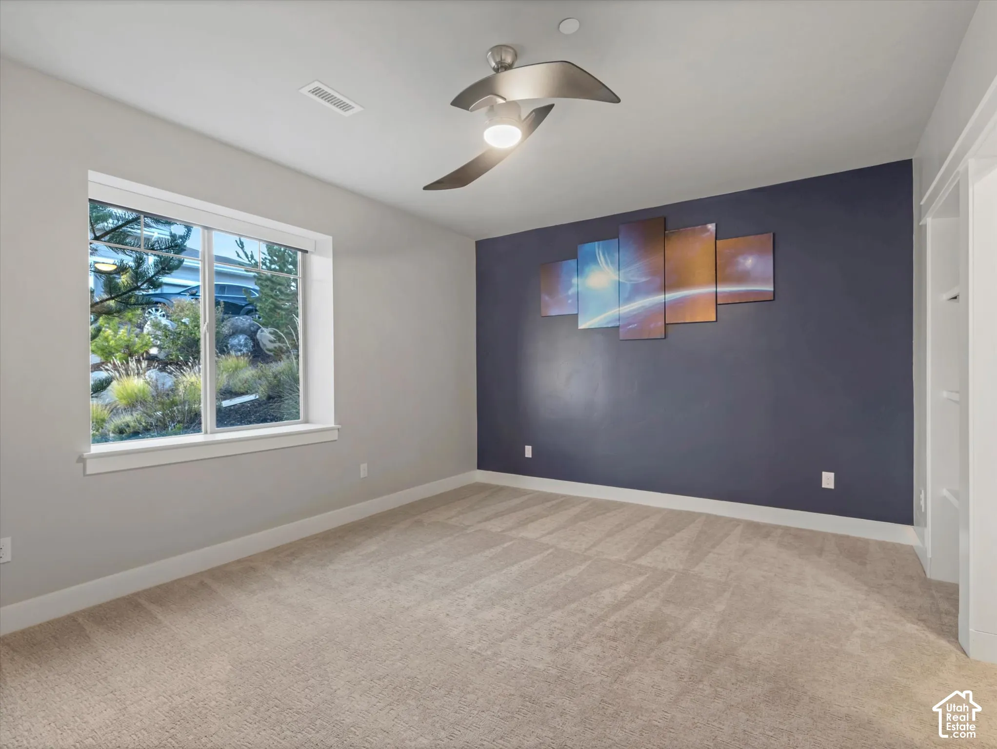 Room featuring carpeted flooring, a dark-toned accent wall, a multi-blade ceiling fan with integrated lighting, and a large window with exterior landscaping
